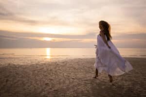 A woman in white dress walking on the beach by the sea in sunset.