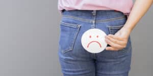 Cropped shot of a woman suffers from hemorrhoids and holds a paper with sad face over light gray background. Hemorrhoid problem concept, copy space.